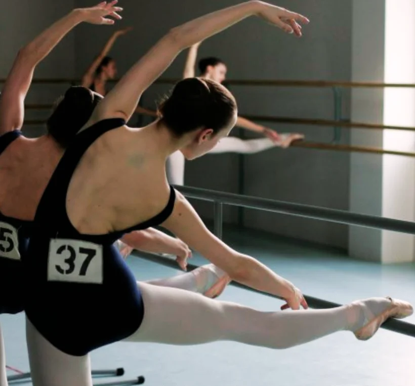 Allieve di danza classica in body blu con numero, allungano il braccio alla sbarra durante la lezione
