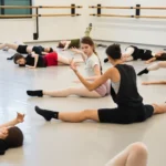 Coaching individuale: perché una lezione su misura fa la differenza Insegnante mostra un gesto a un'allieva mentre altri ballerini sono distesi in sala