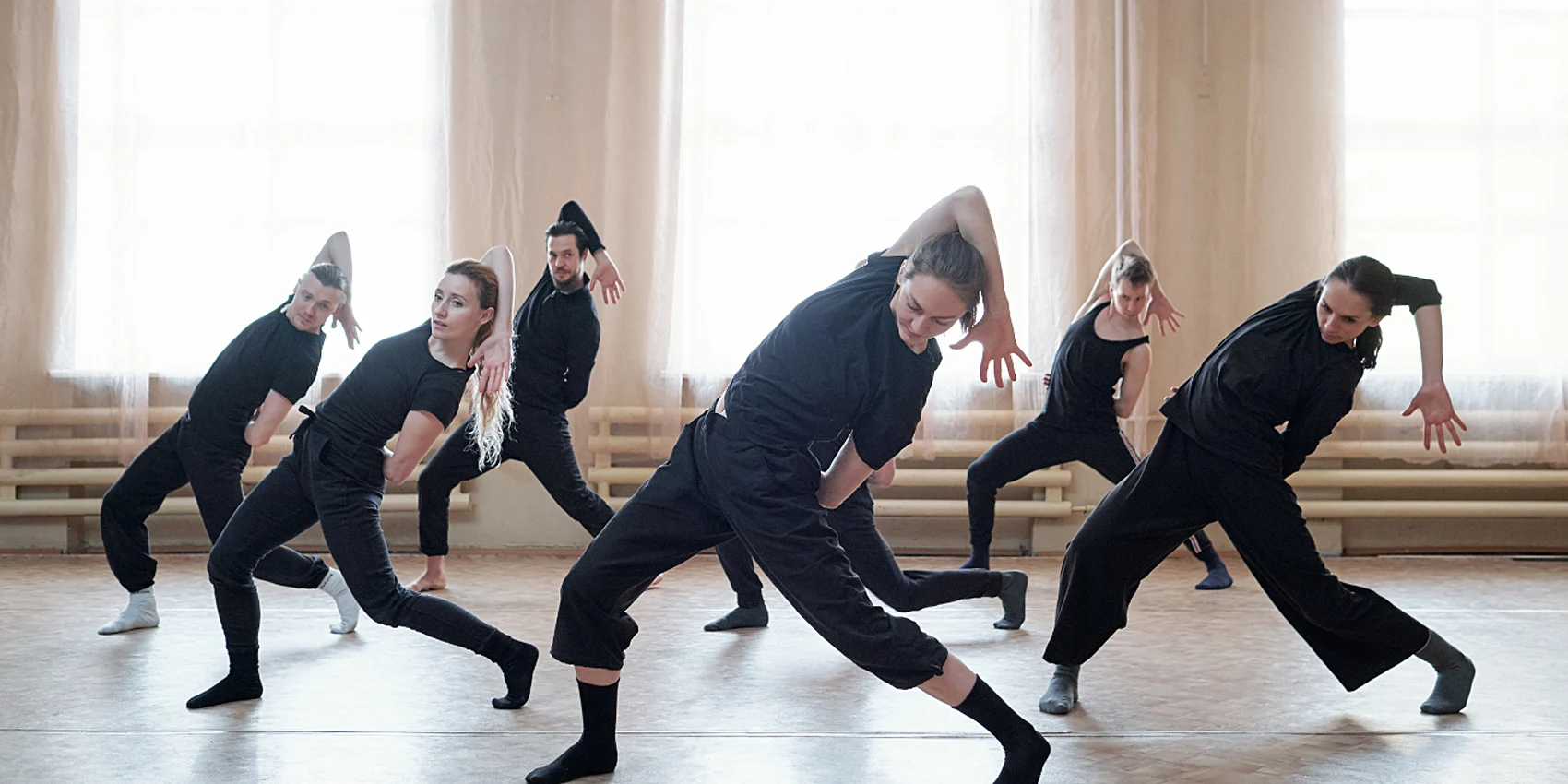 Gruppo di danzatori in abiti neri in posa dinamica in una sala prove luminosa con grandi finestre