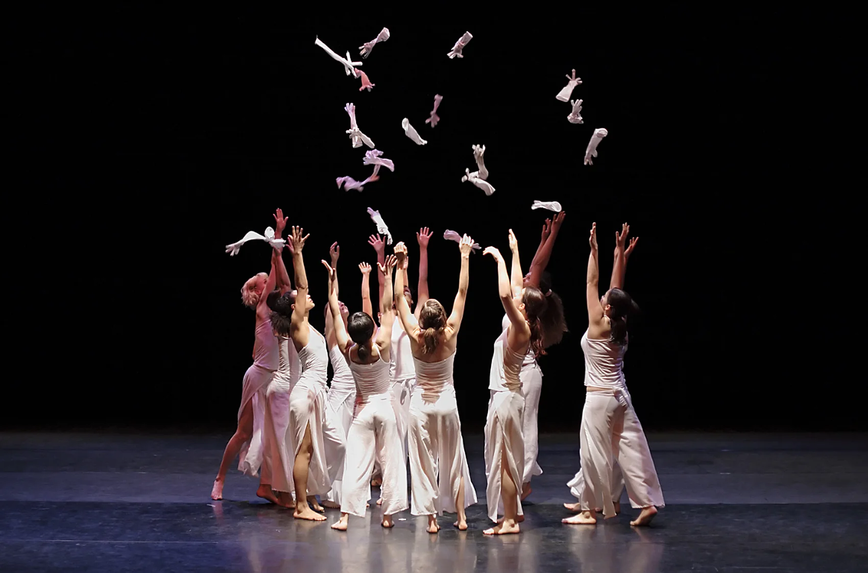Gruppo di danzatrici in abiti bianchi che lanciano guanti in aria sul palcoscenico nero