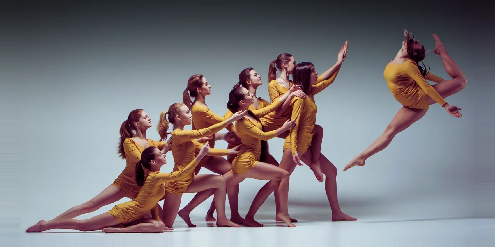 Gruppo di danzatrici in body giallo senape che danzano in sequenza su sfondo studio grigio azzurro